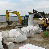 Relleno con arena local de los sacos SoilTain con la ayuda de una excavadora mientras una cargadora de ruedas sostiene el saco de arena prefabricado.
