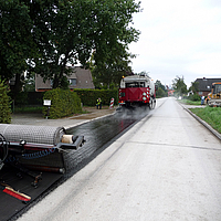 Una máquina de término avanza, seguida de la aplicación del refuerzo de asfalto SamiGrid® en la carretera